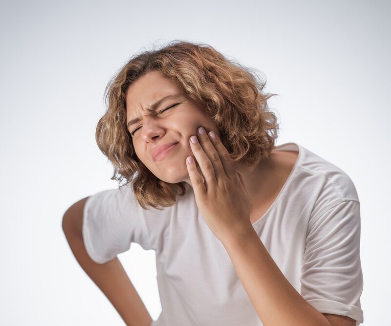 Young woman grimaces in pain while pressing her cheek, showing clear signs of oral discomfort due to tooth or gum issues
