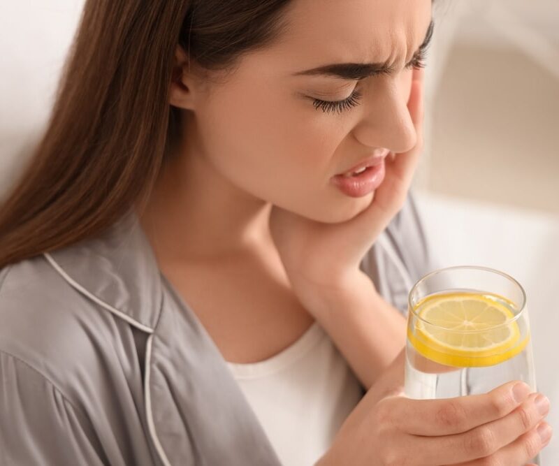 Young woman in pajamas with lemon water suffering from toothache at home, closeup