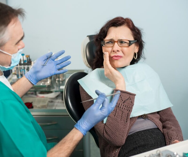 Woman feeling toothache, touching cheek with hand at dental clinic. Senior dentist trying to help
